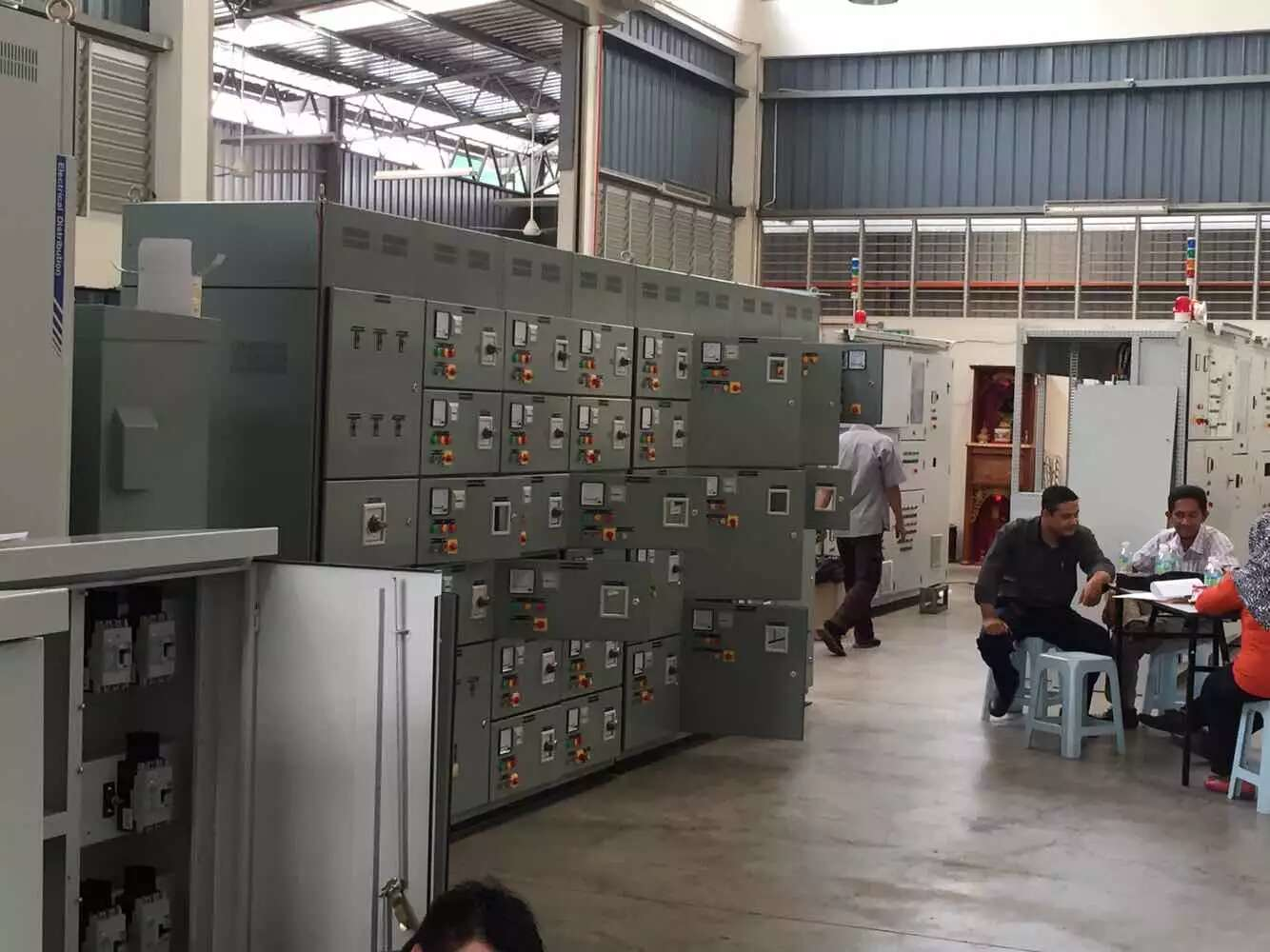 Electrical control panels in an industrial setting with workers at a table; gray and white tones.