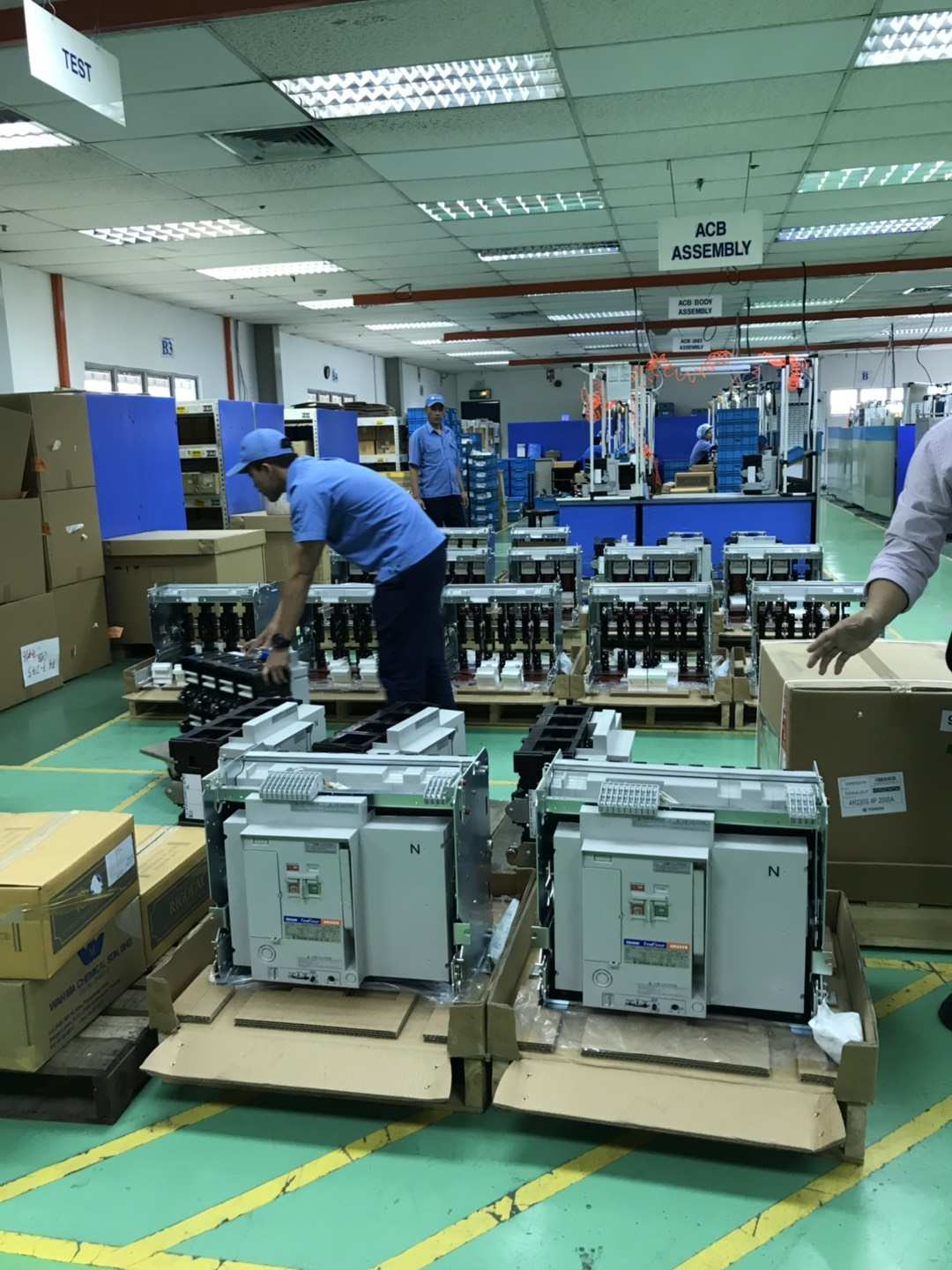 Factory workers assembling electrical equipment on a green floor. Men in blue uniforms work around large circuit breakers.