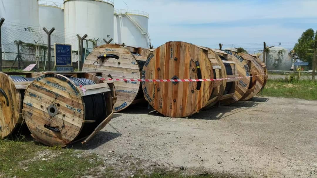 Wooden cable spools on a gravel surface, with storage tanks and a fence in the background on a sunny day.