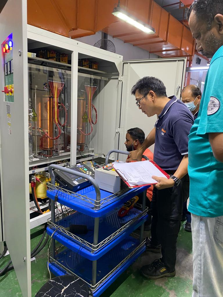 Men inspecting machinery in a facility; one points to a control panel, others observe.