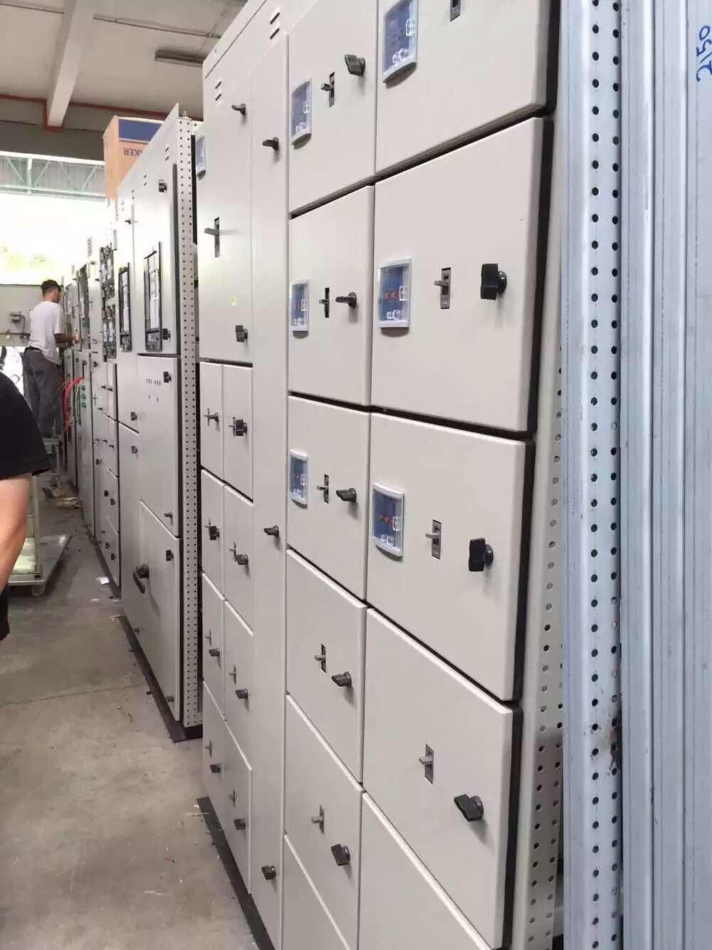 A row of light-colored electrical control panels with black handles and digital displays in a factory.