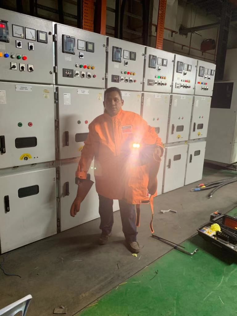 Man in orange work suit stands in front of electrical control panels. Bright light source.