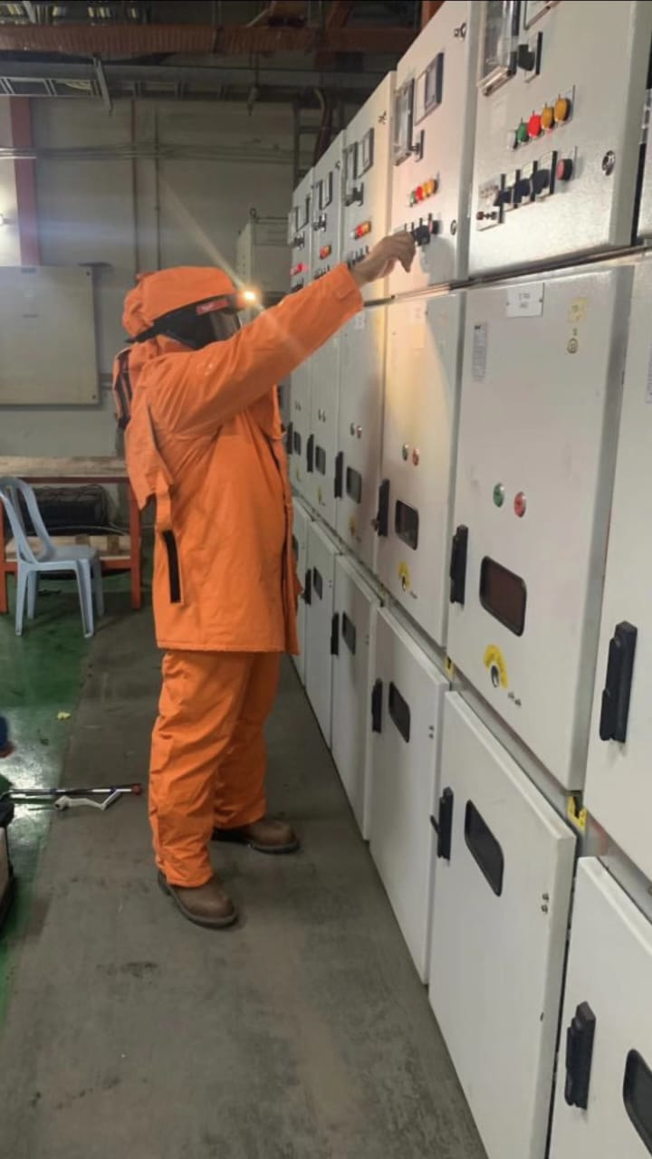Person in orange safety suit points at electrical panel. Industrial setting.