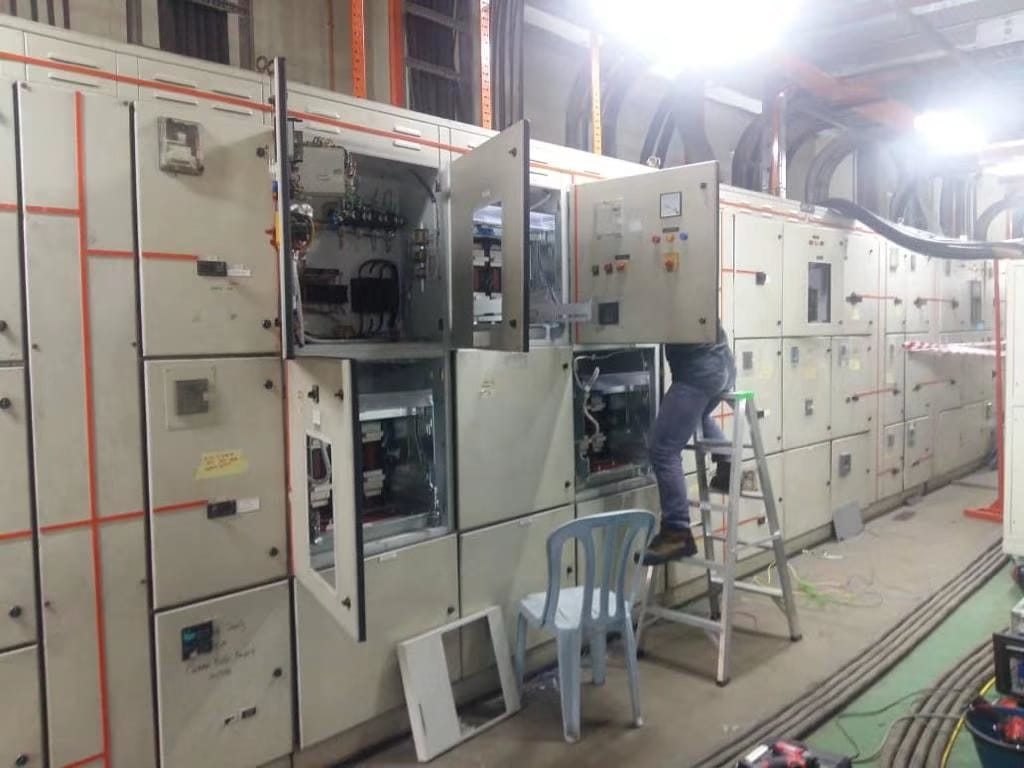 Person on a ladder working on electrical panels in an industrial setting; multiple panels visible.