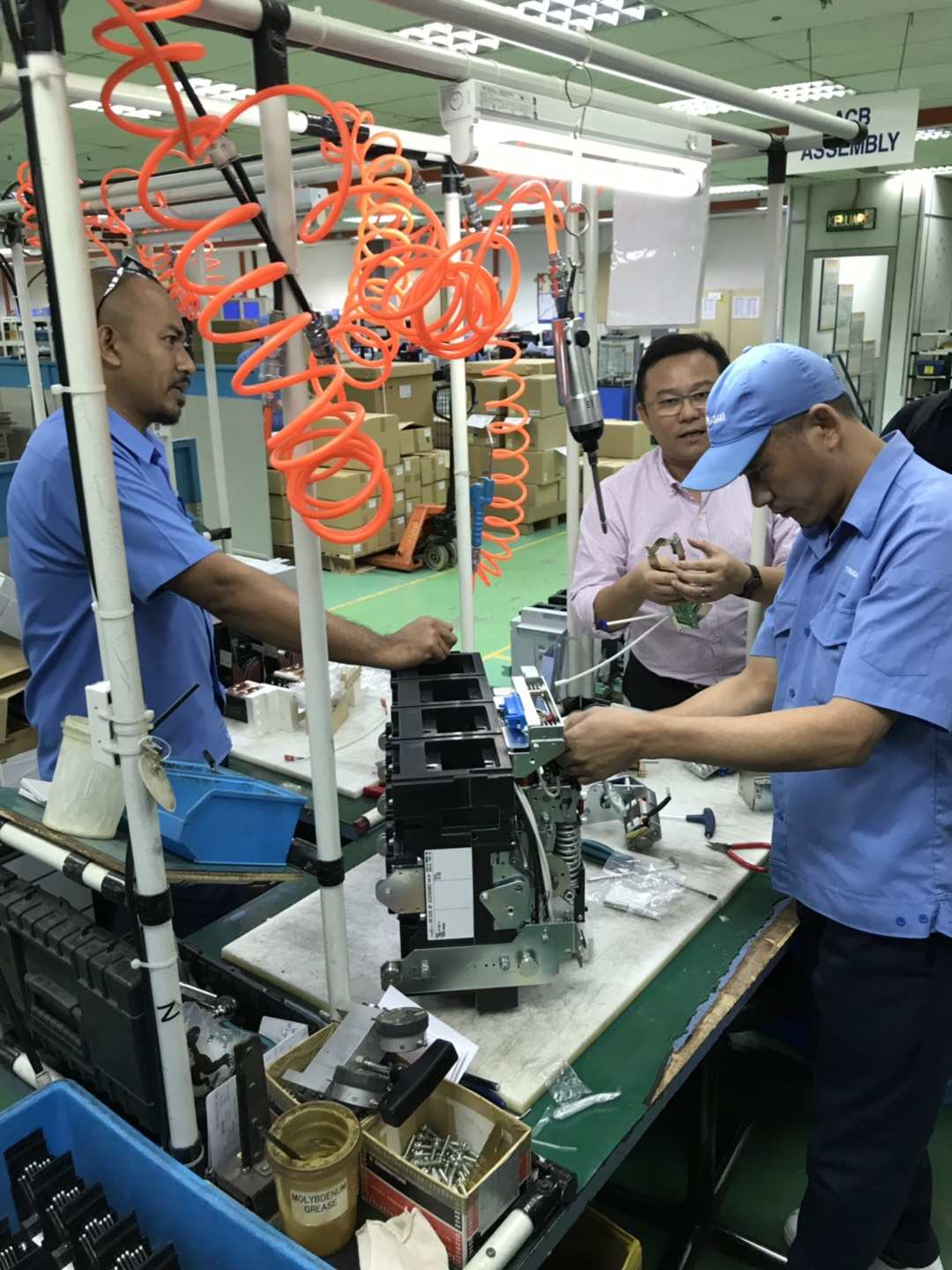 Workers assemble a device on a factory line. One man is working, another observes, and a third stands nearby.