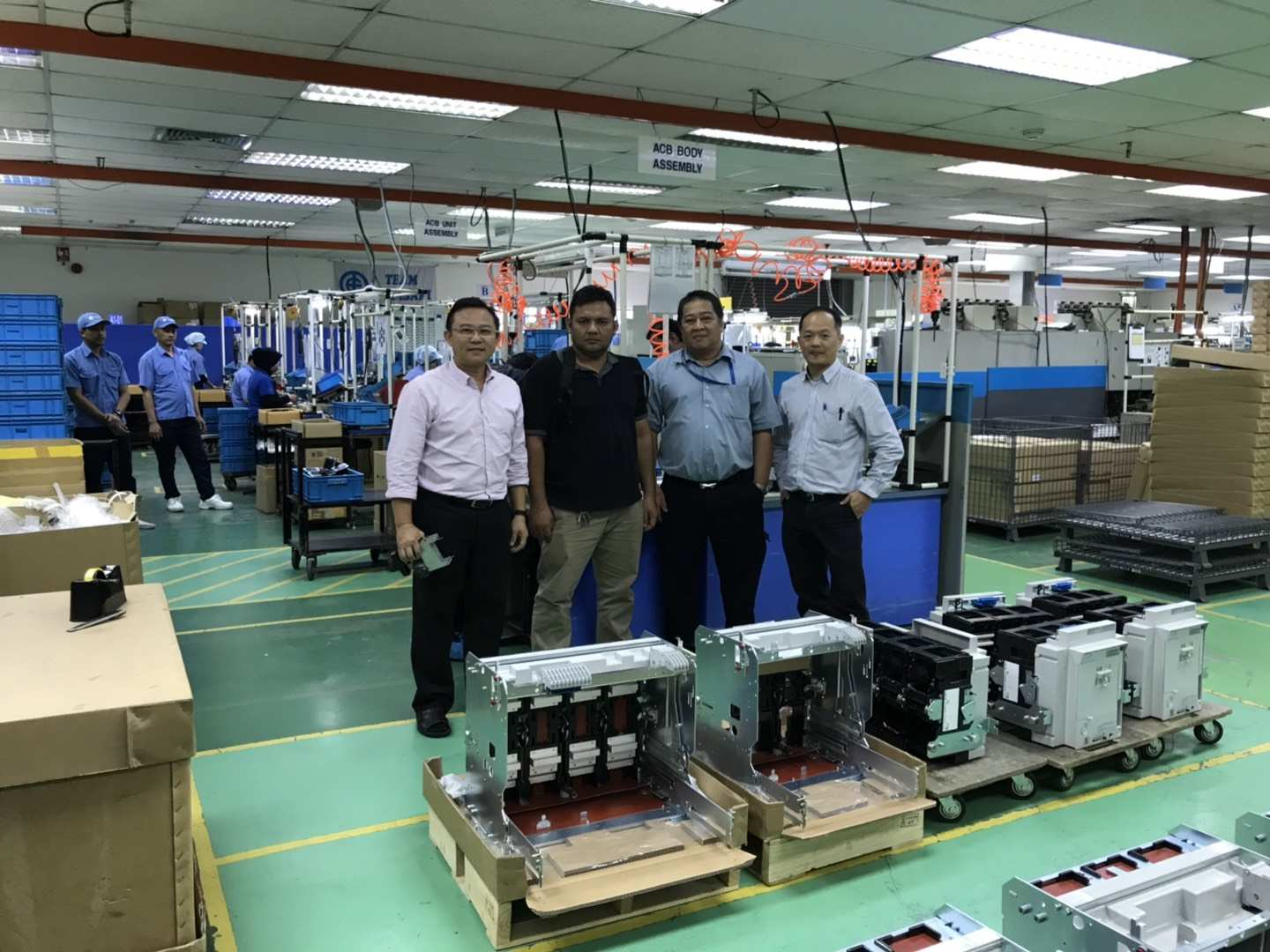 Four men pose in a factory with assembled electrical equipment; other workers in the background.