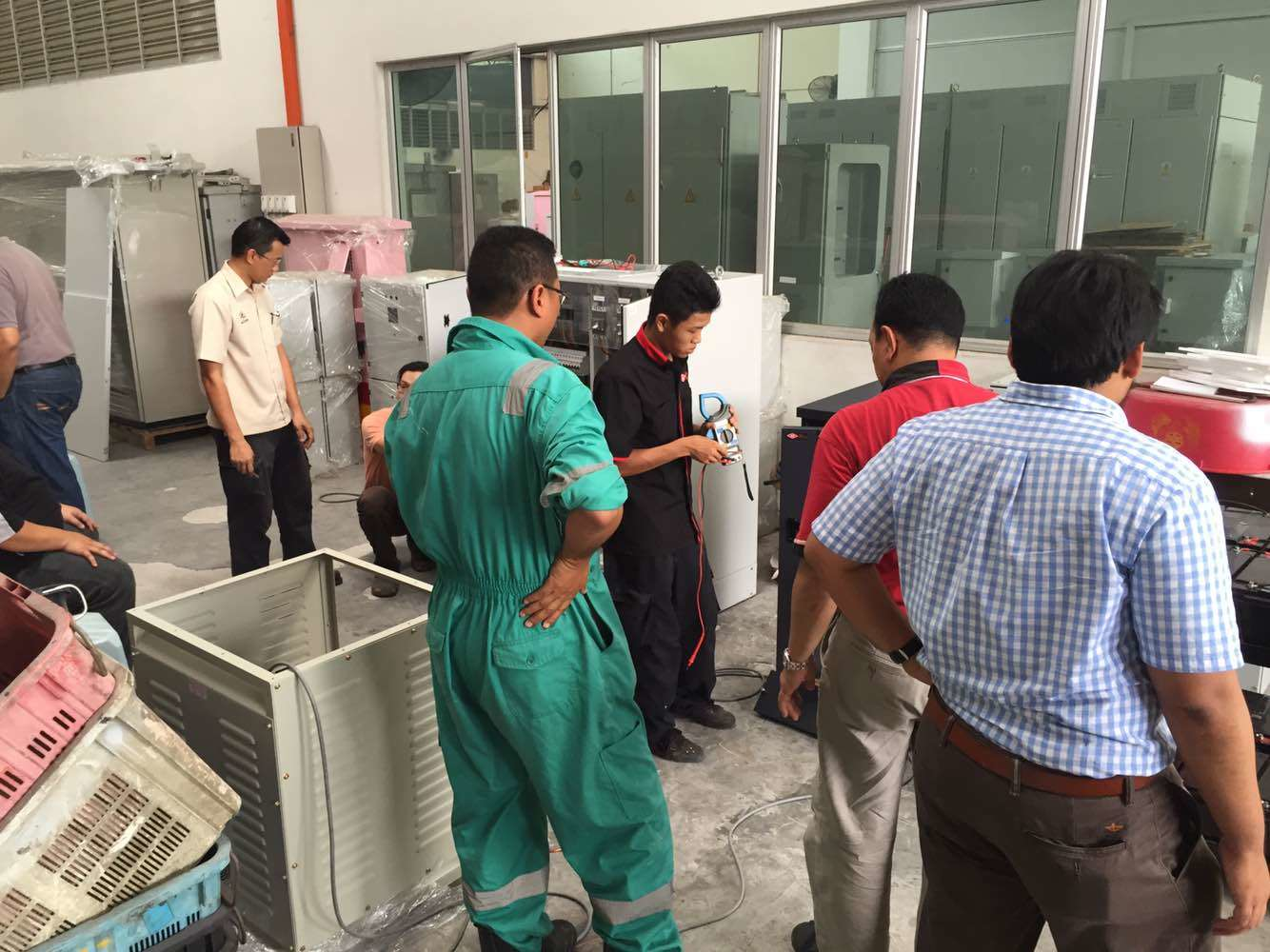 A group of men in a workshop, observing and working on machinery. Green overalls, red and blue shirts.