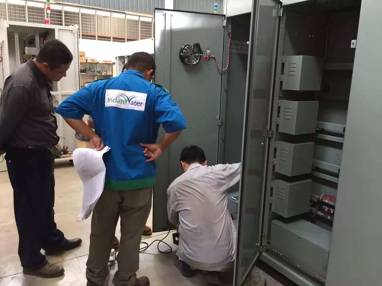 Three men in a factory setting, working on electrical equipment within a gray cabinet.