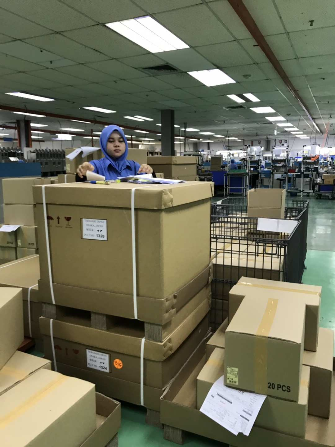 Woman in hijab working at a factory, surrounded by stacked cardboard boxes and manufacturing equipment.