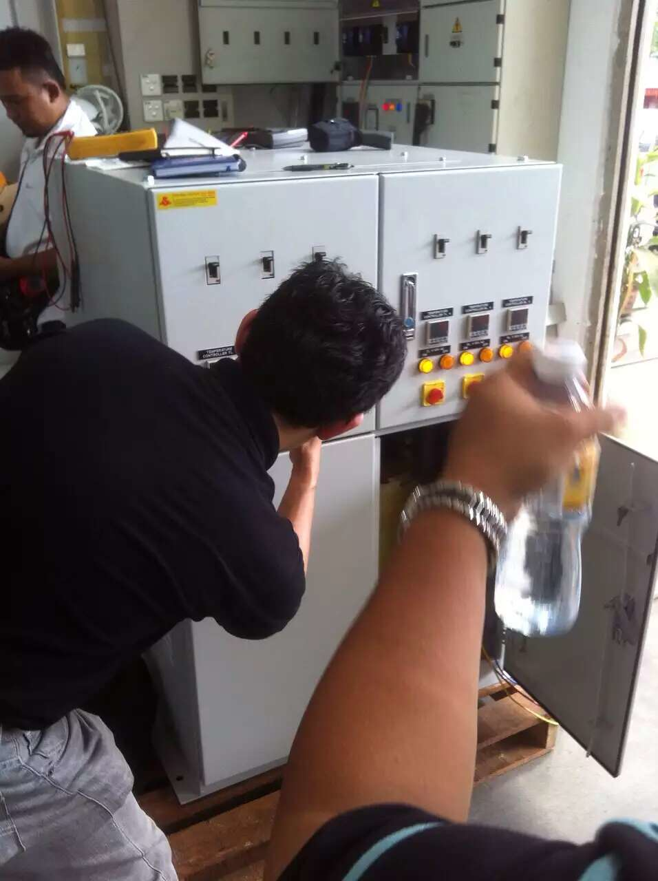 Two men working on a gray electrical panel outdoors; one leans inside, another holds a water bottle.