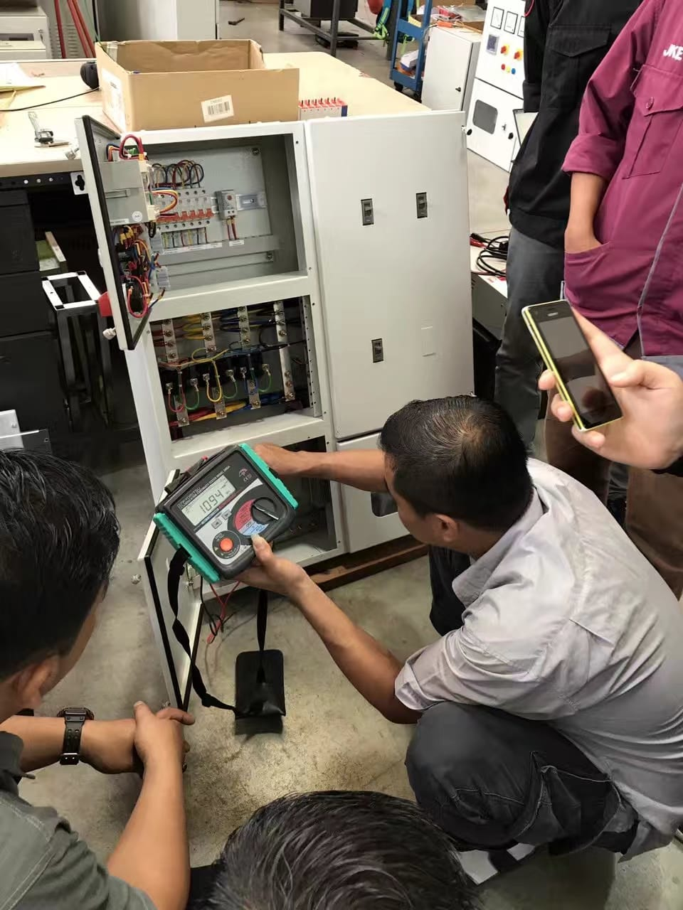 A man in a gray shirt checks electrical panel with a multimeter while others watch.