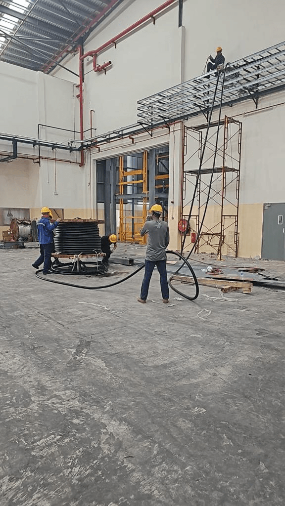 Construction workers installing cables in a warehouse. One on a scaffold, others on the floor.