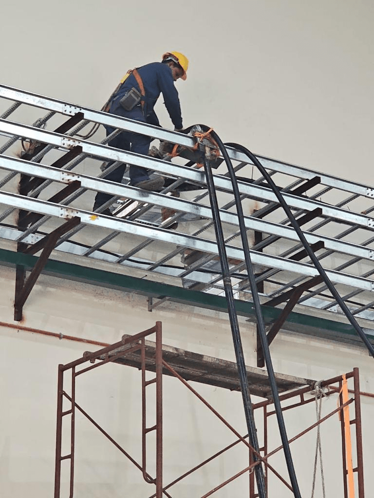 Construction worker in yellow hardhat on scaffolding, installing cables along a metal cable tray.