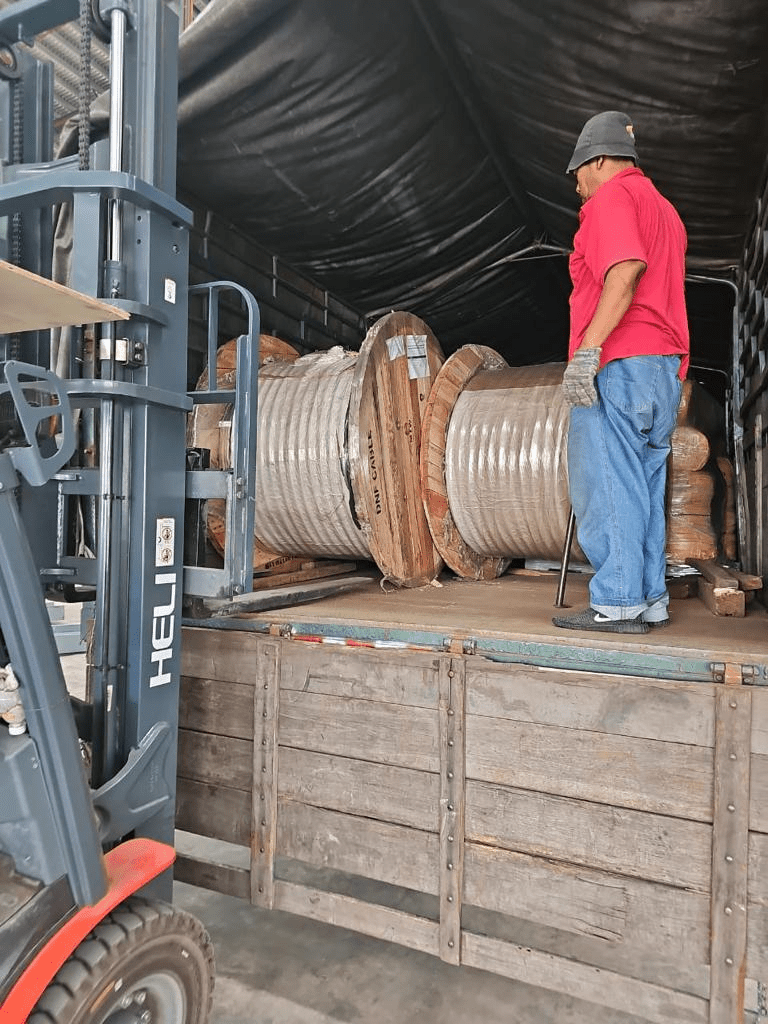 Forklift loading large spools of cable into a truck, observed by a worker in a red shirt.