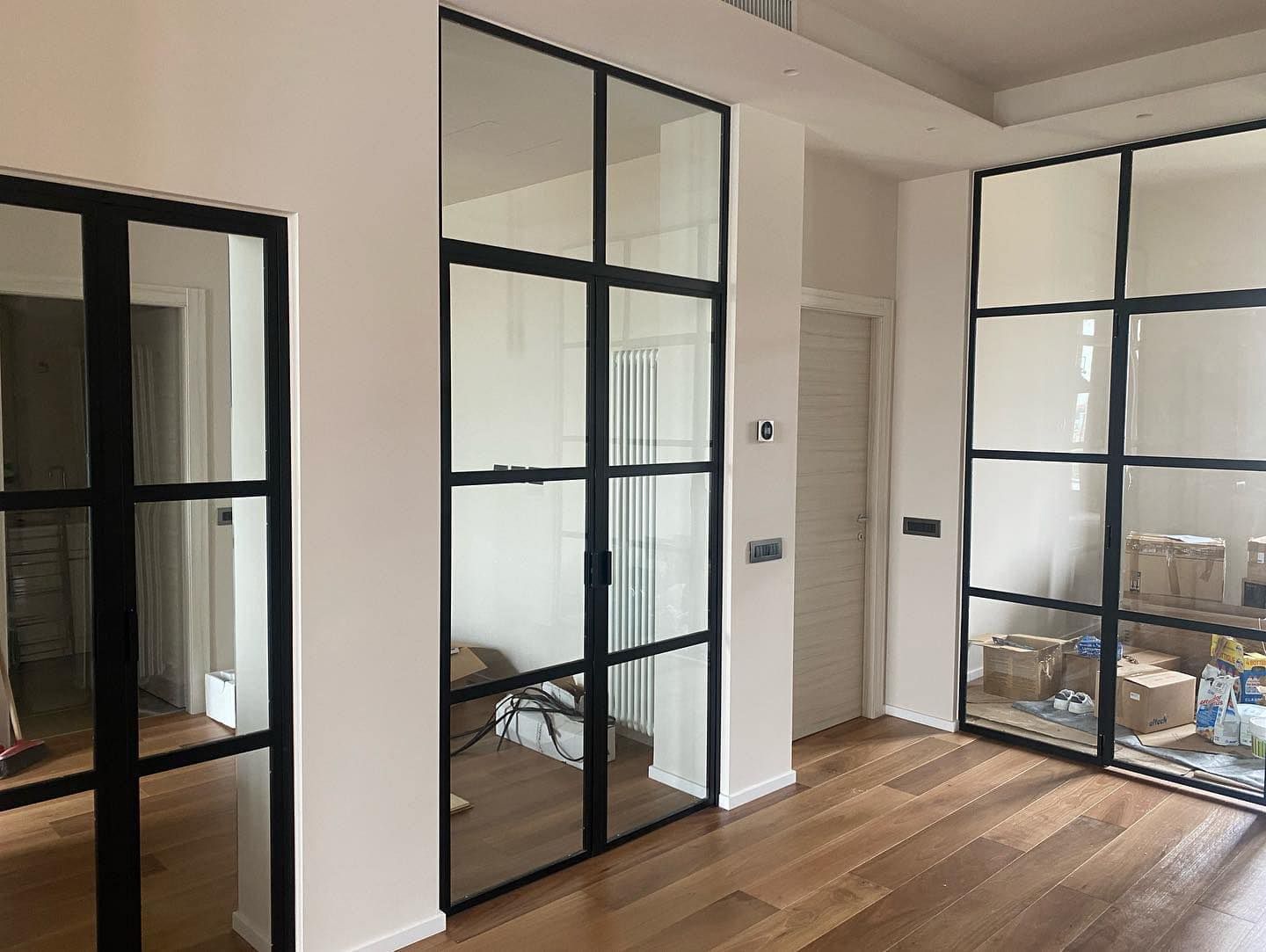 Interno con porte in vetro con cornice nera e pavimenti in legno, che conducono a diverse stanze con pareti bianche.