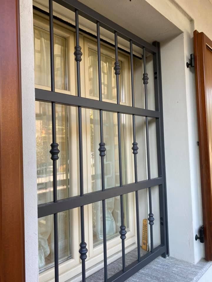 Inferriate di sicurezza in metallo scuro su una finestra con telaio beige e persiana marrone.