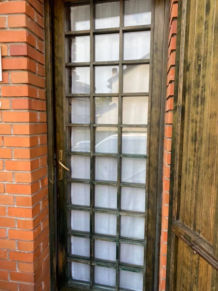 Porta in legno scuro con pannelli di vetro a griglia, accanto a un muro di mattoni e un pannello di legno.