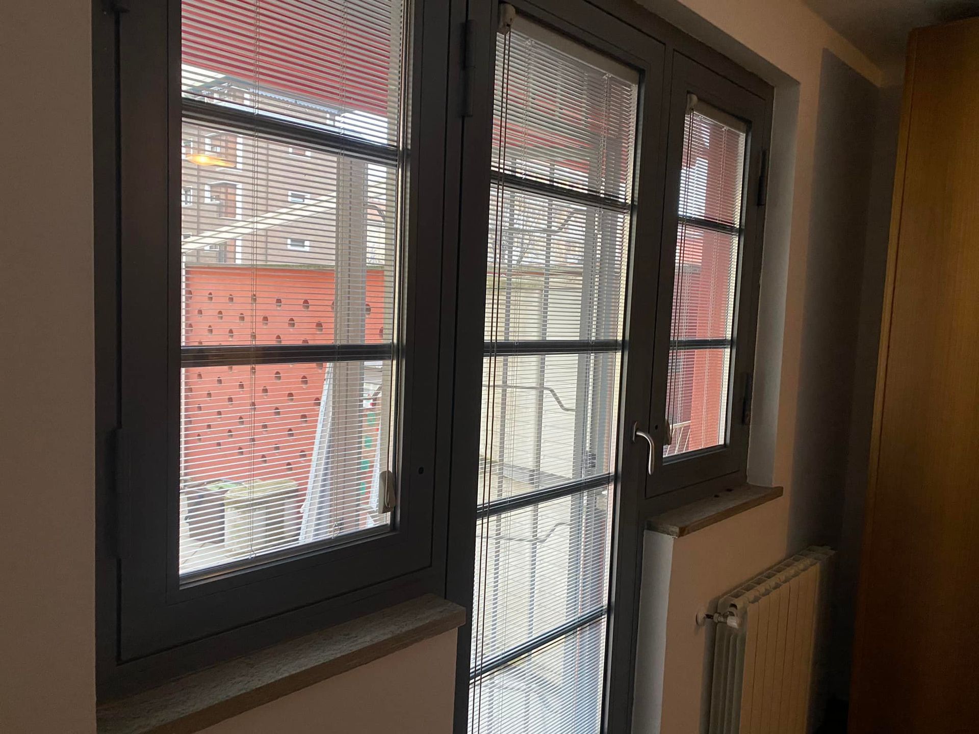 Finestra grigia con vetro strutturato, che si affaccia su un edificio in mattoni rossi e un termosifone.