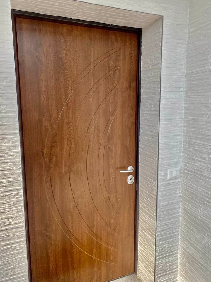 Porta in legno con scanalature curve e maniglia argentata, inserita in una parete chiara e strutturata.