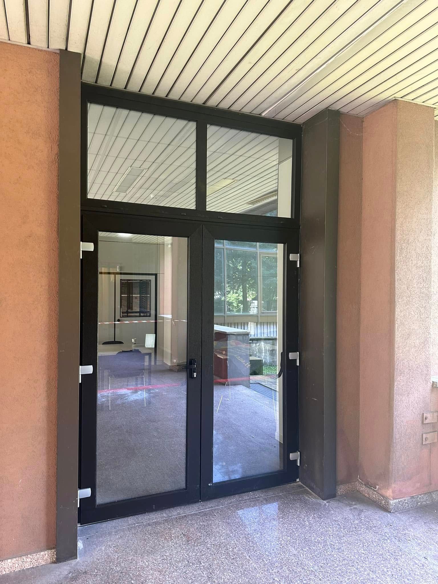 Porta in vetro con cornice nera e pannelli superiori, tra pilastri marroni. L'interno è visibile attraverso la porta.