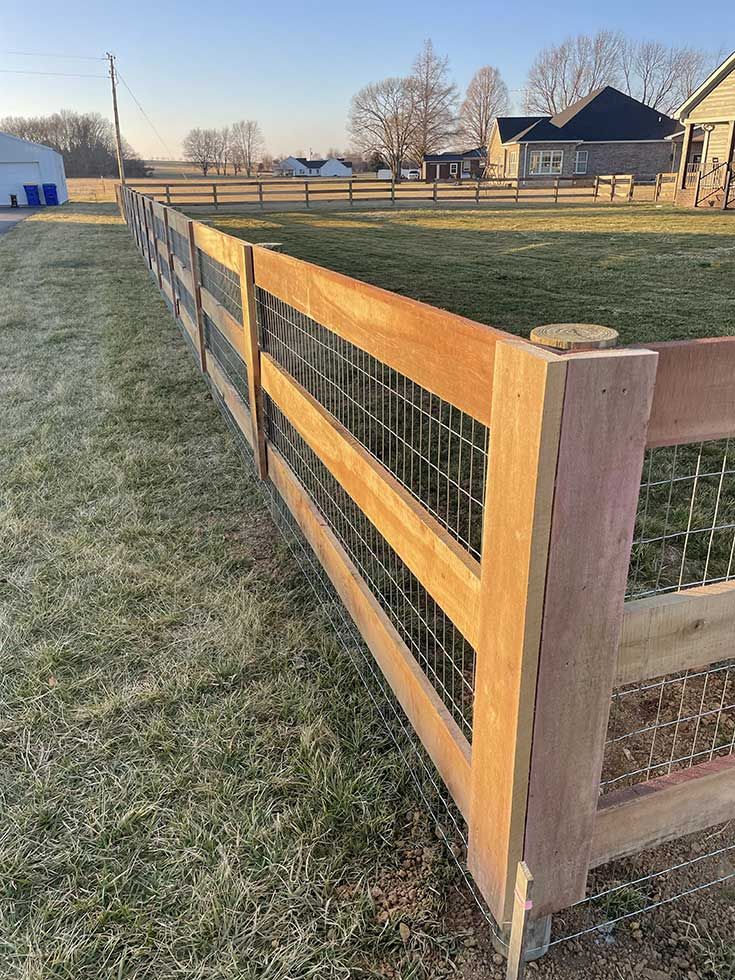 Wooden fence with wire mesh panels in a grassy field, houses and trees in the background.