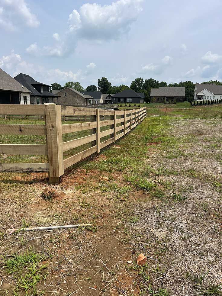 Wooden fence in a field, houses in the background, blue sky with clouds.