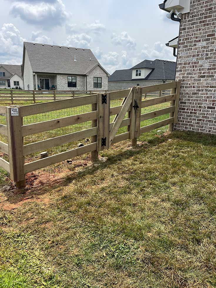 Wooden fence with a gate next to a brick house on a grassy yard.