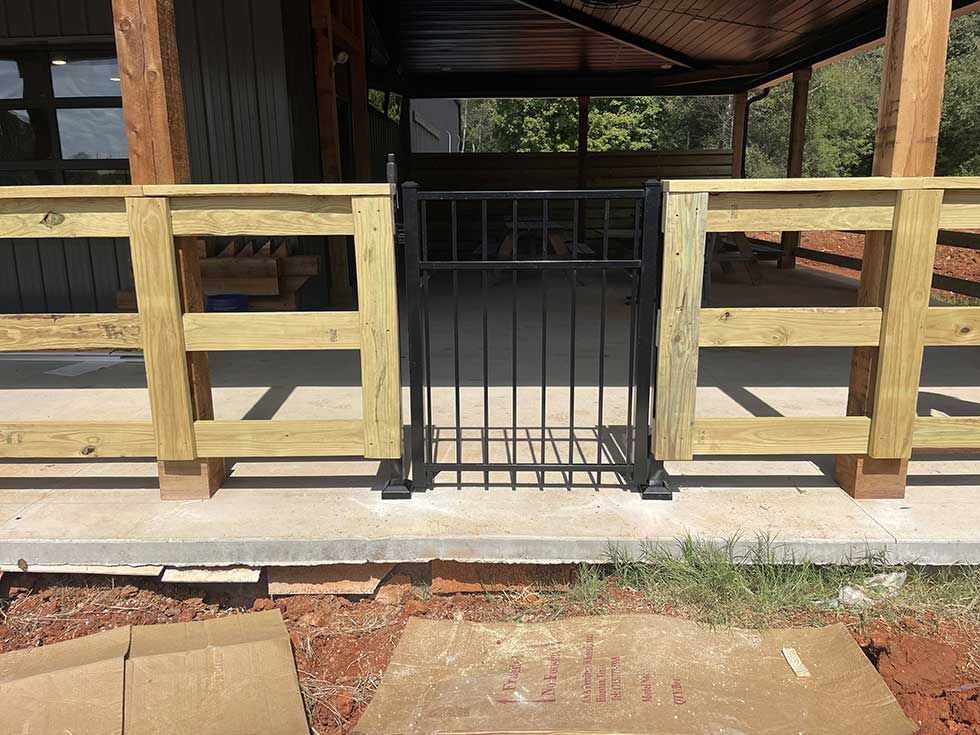 Black metal gate in a light brown wooden fence on a concrete patio, leading to a covered porch.