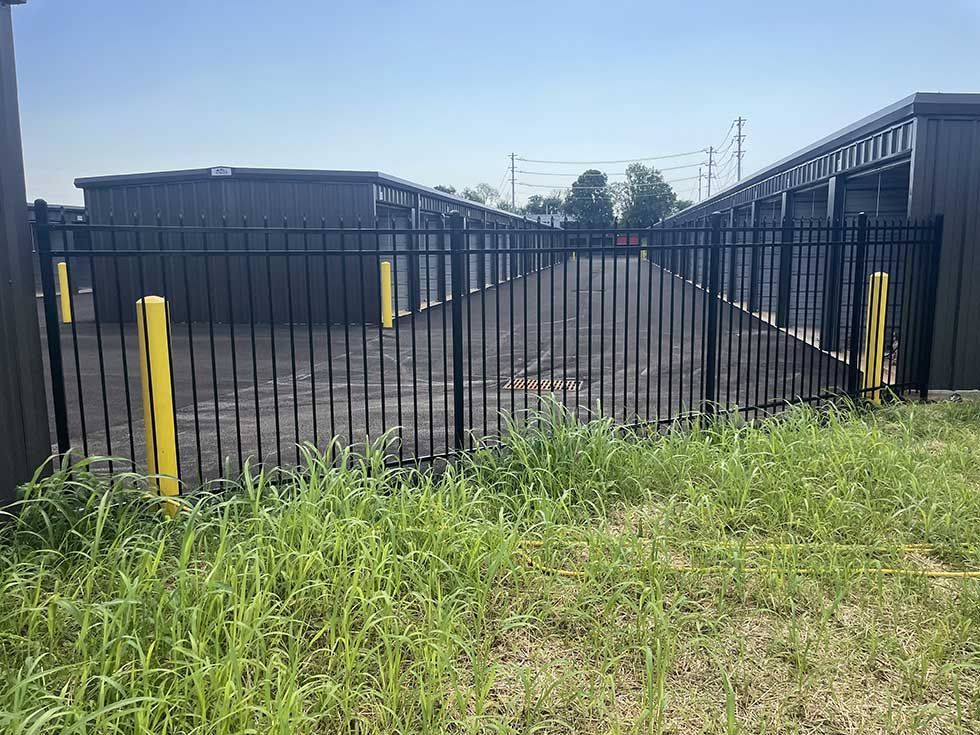 Black metal fence with storage units in the background. Green grass and yellow posts.