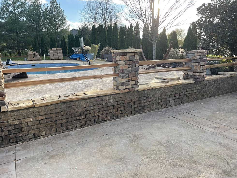 Stone fence with wooden rails and stone pillars bordering a concrete patio, with a pool in the background.
