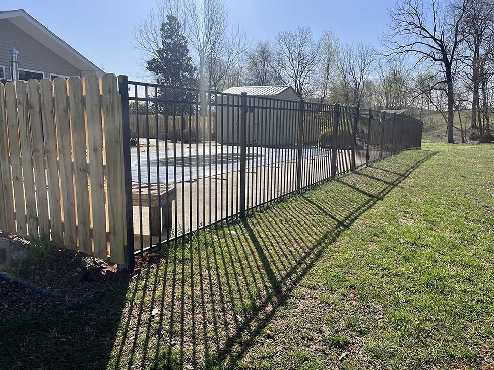 Black metal fence surrounding a swimming pool and partially bordering a wooden fence on a sunny day.