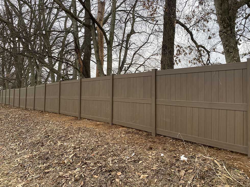 A brown vinyl fence extends along a wooded area, set in front of bare trees and fallen leaves.