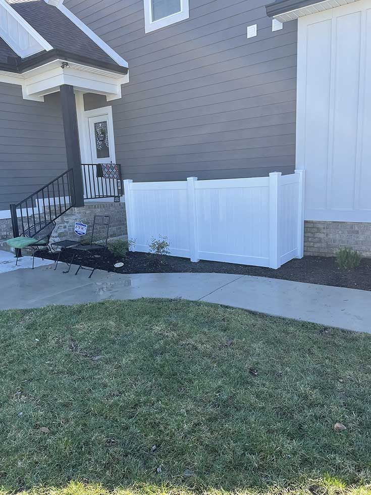 White fence encloses an area next to a house with a gray exterior, a door, and a walkway.