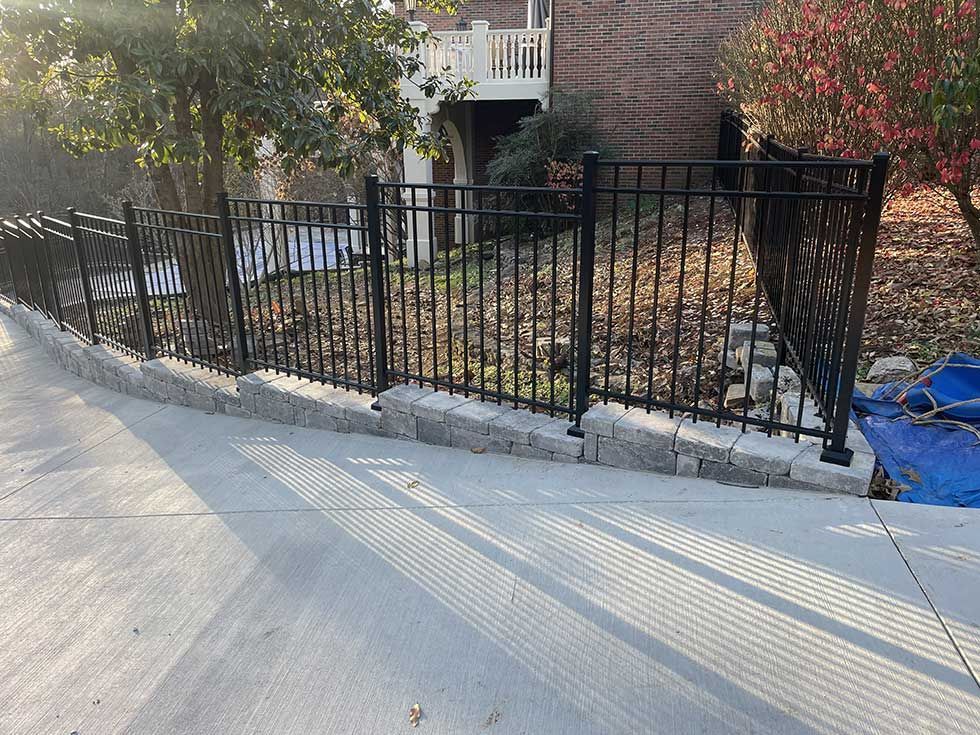 Black metal fence atop a stone retaining wall bordering a concrete walkway. Trees and a brick building in the background.