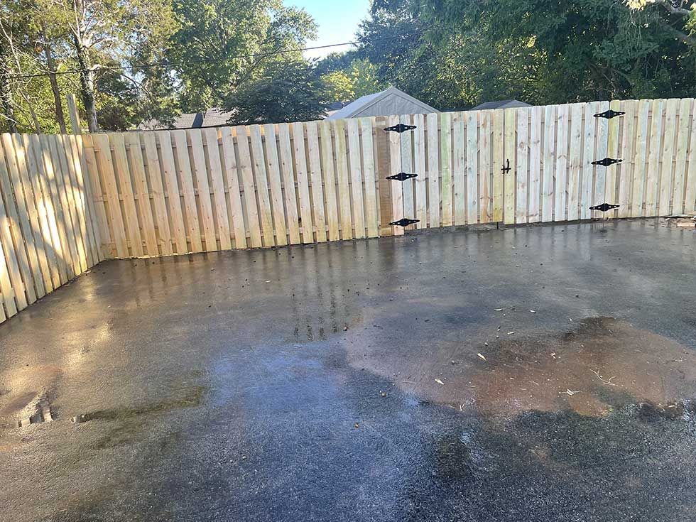 Wooden fence with gate in an asphalt-paved area, with sunlight and trees in the background.