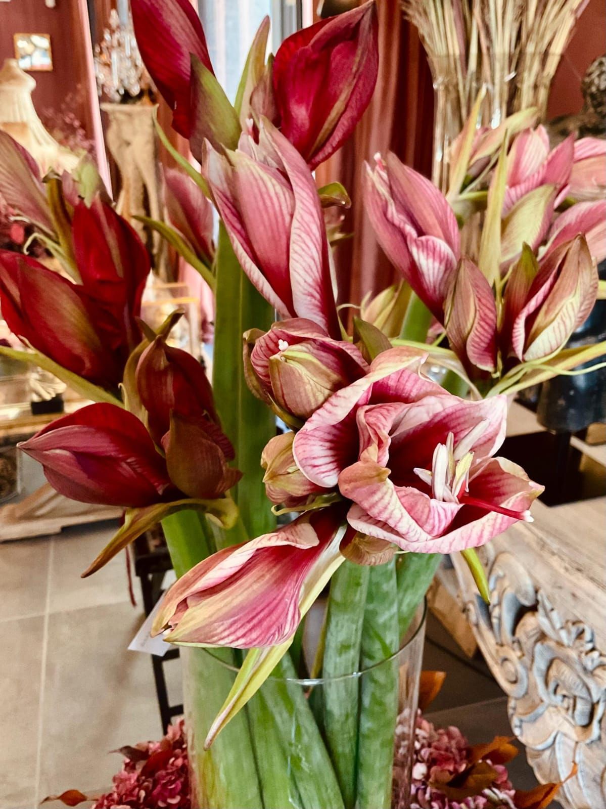 Fiori di amarilli rossi e rosa in un vaso di vetro, con steli e boccioli verdi in un ambiente interno.