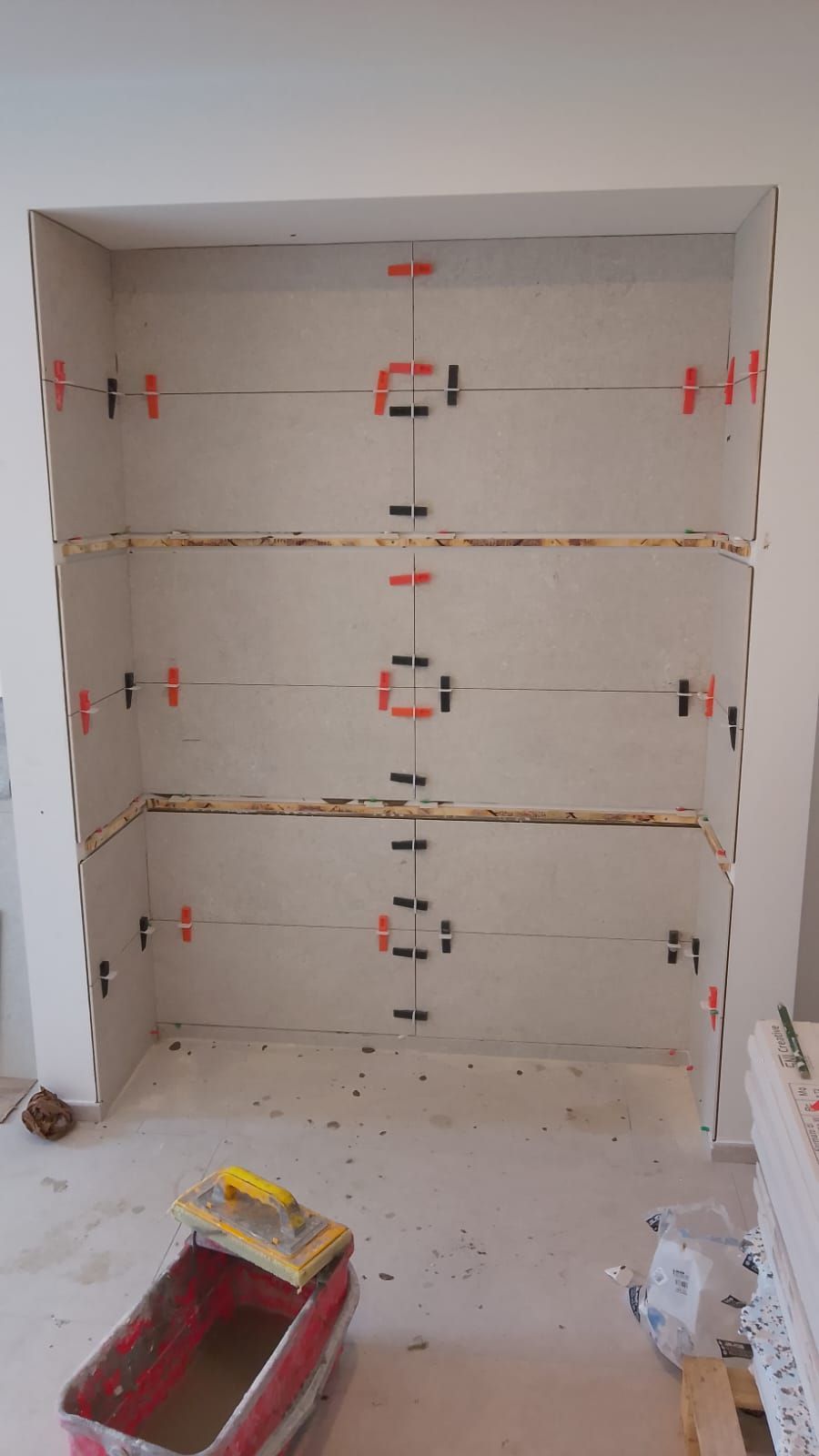 Grandi piastrelle da parete di colore chiaro, installate in una nicchia incassata utilizzando un sistema di livellamento in plastica rossa e nera.