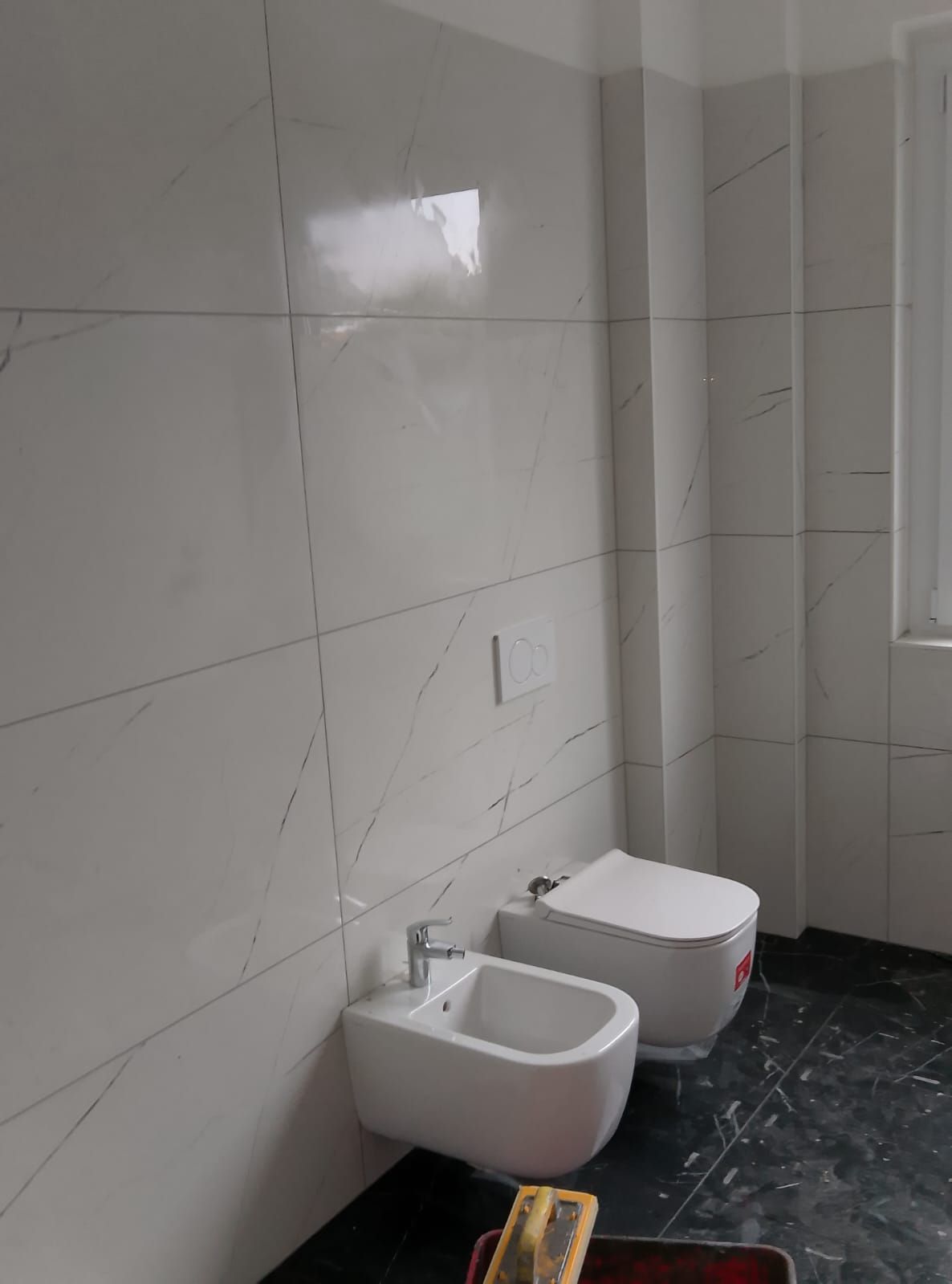 Un bidet e un WC sospesi di colore bianco in un bagno moderno con piastrelle a parete effetto marmo chiaro e piastrelle a pavimento scure.