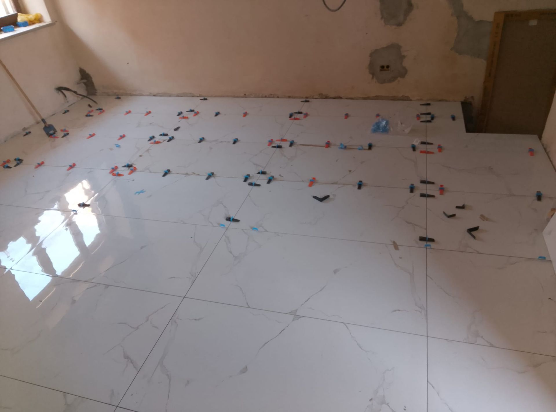 Un pavimento parzialmente piastrellato con piastrelle effetto marmo bianco, distanziatori e clip di fissaggio in una stanza non rifinita.
