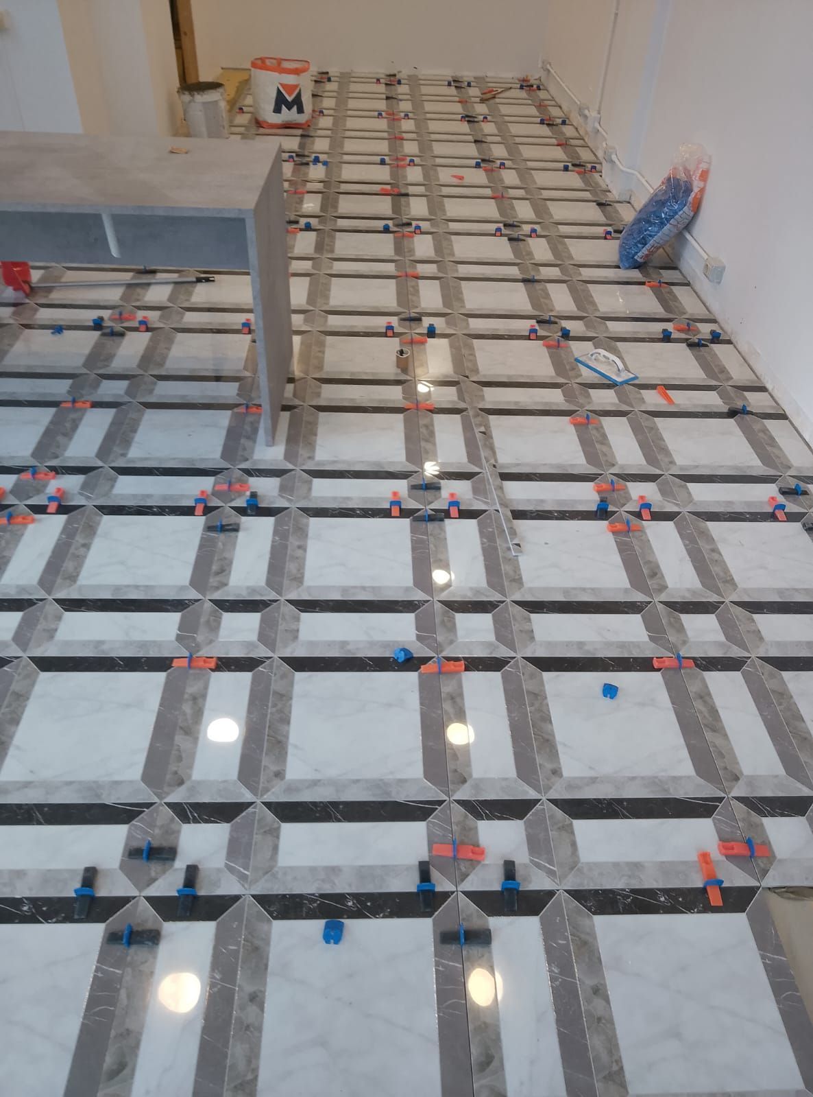 Una stanza con pavimento piastrellato con motivi geometrici bianchi e grigi, in fase di installazione, con distanziatori in plastica colorata.