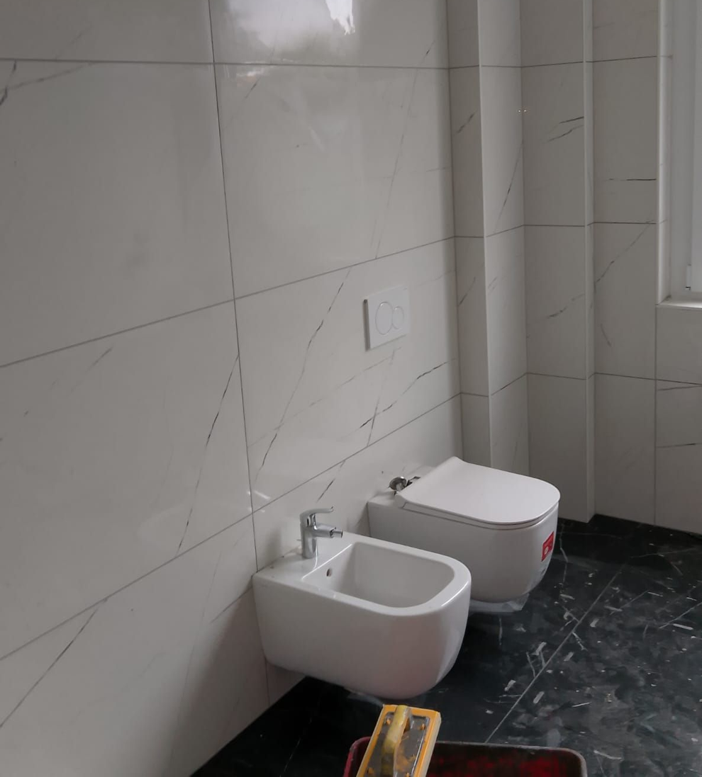Un bidet bianco e un WC sospeso installati su piastrelle chiare effetto marmo in un bagno con pavimento piastrellato scuro.