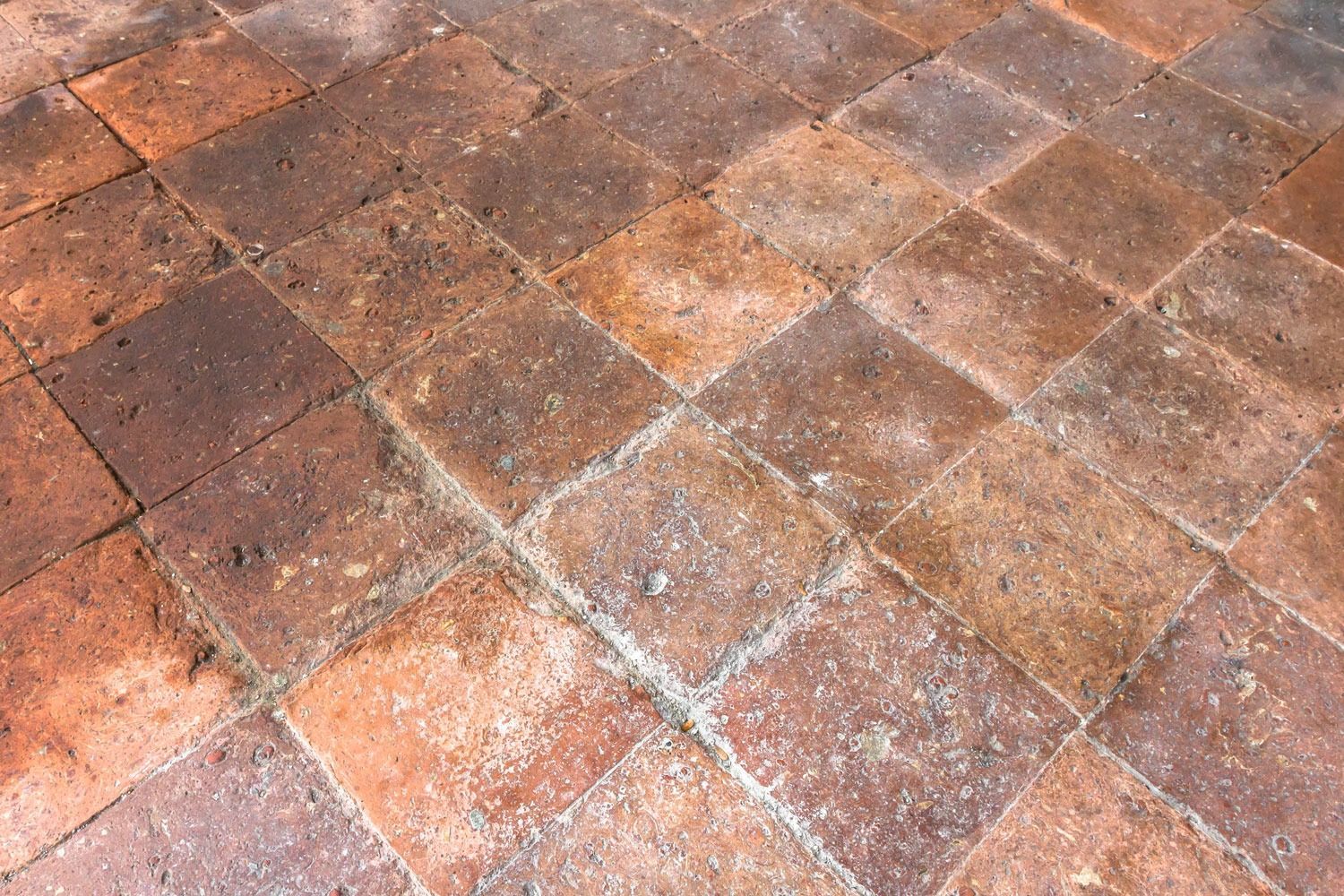 Una superficie di piastrelle quadrate in terracotta di colore marrone rossastro, usurate dagli agenti atmosferici, con fughe a vista e texture variegate.