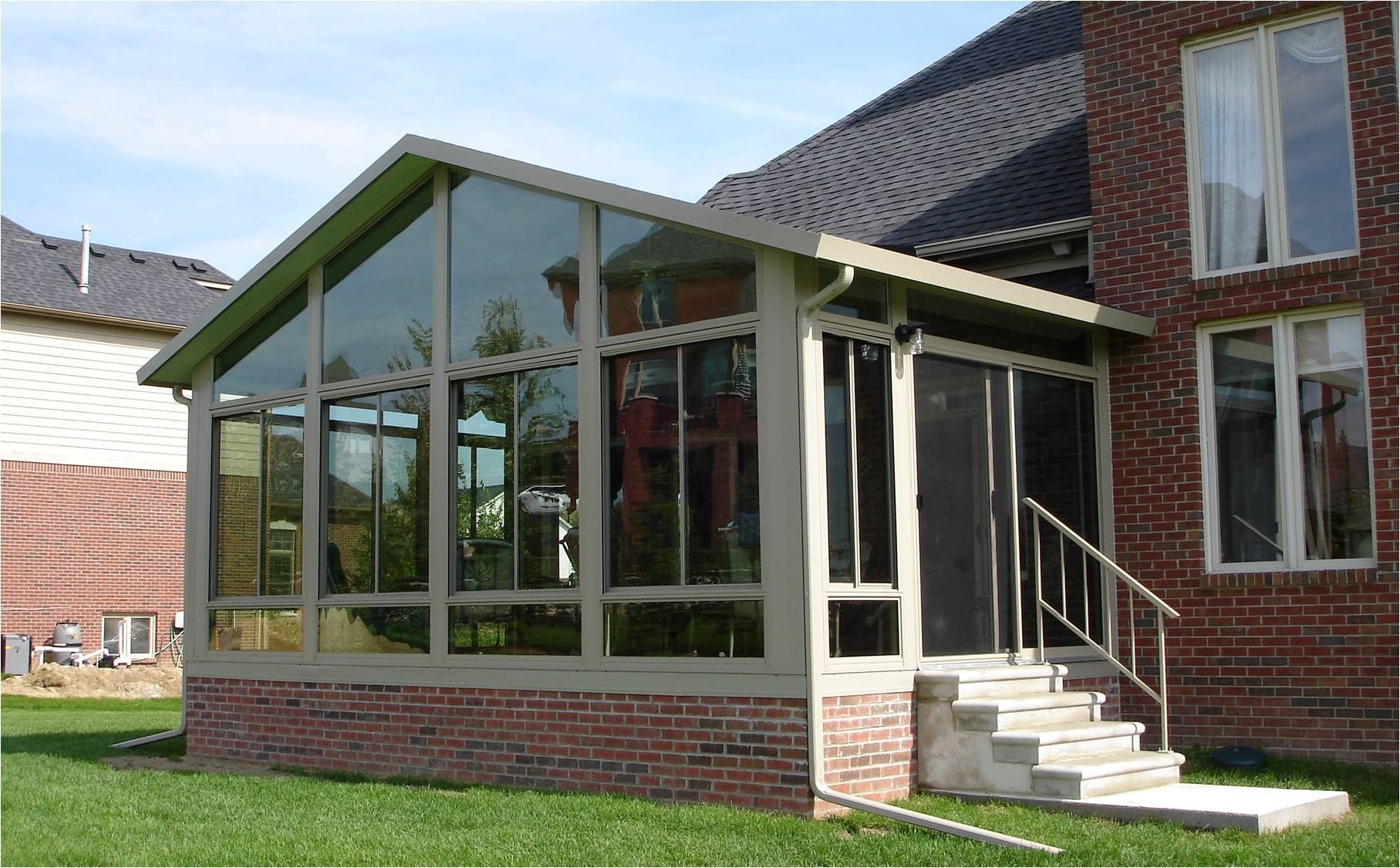 A glass sunroom attached to a brick house. Green grass in foreground; clear sky.