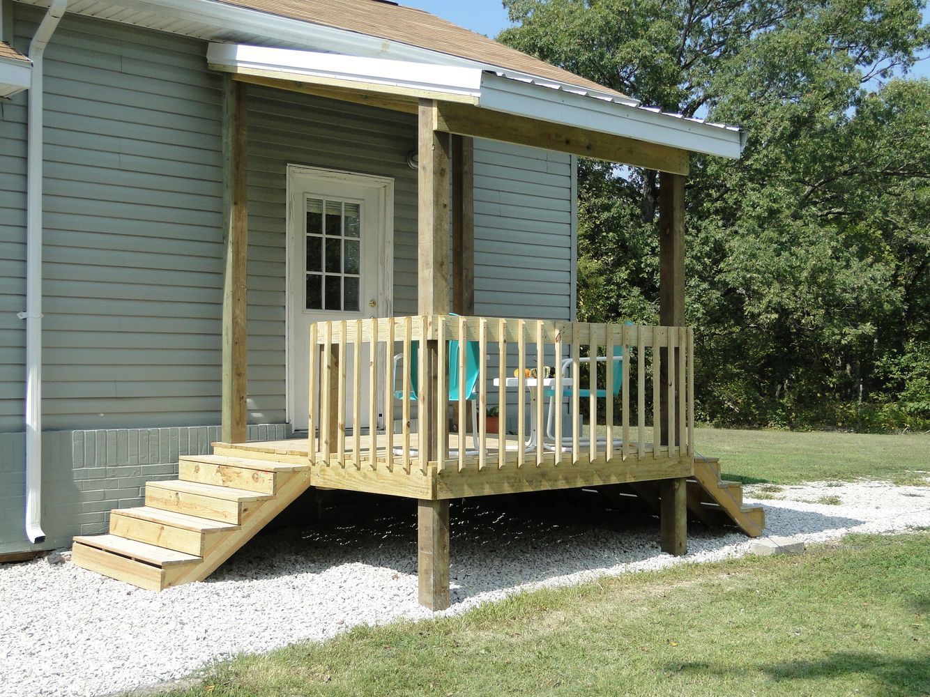 Wooden porch with steps, roof, and railing, attached to a pale blue house with white gravel path and lawn.