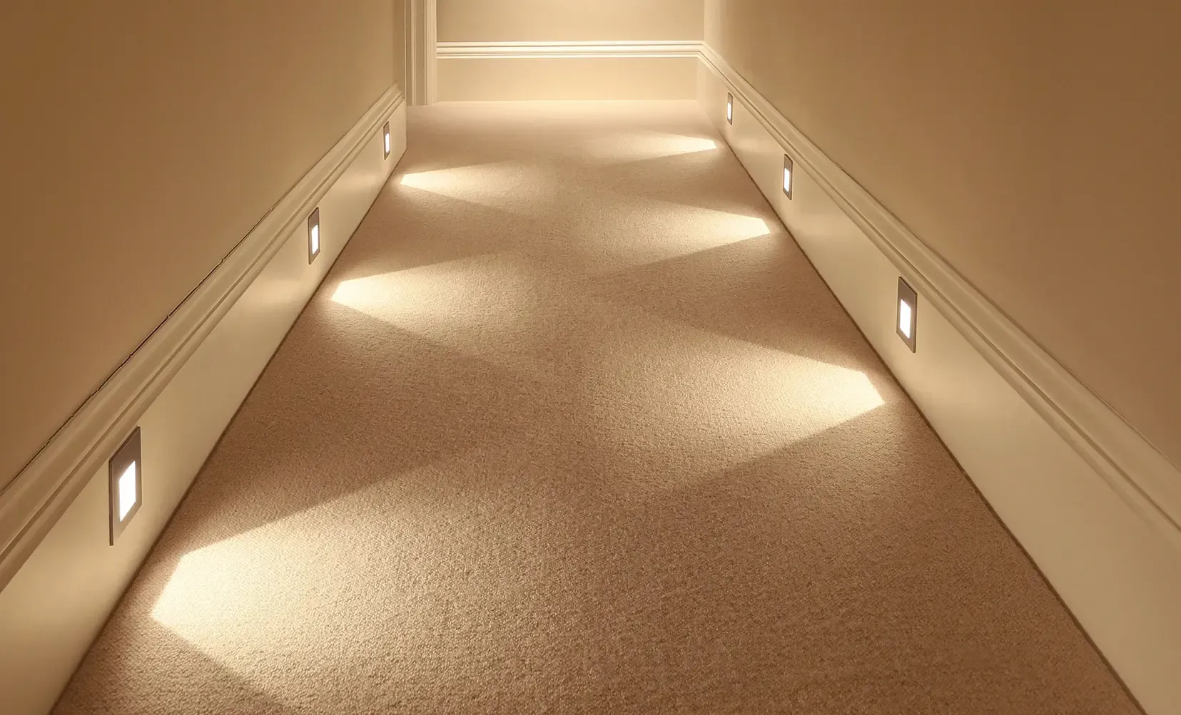A hallway with recessed lights illuminating a tan carpet and off-white walls.