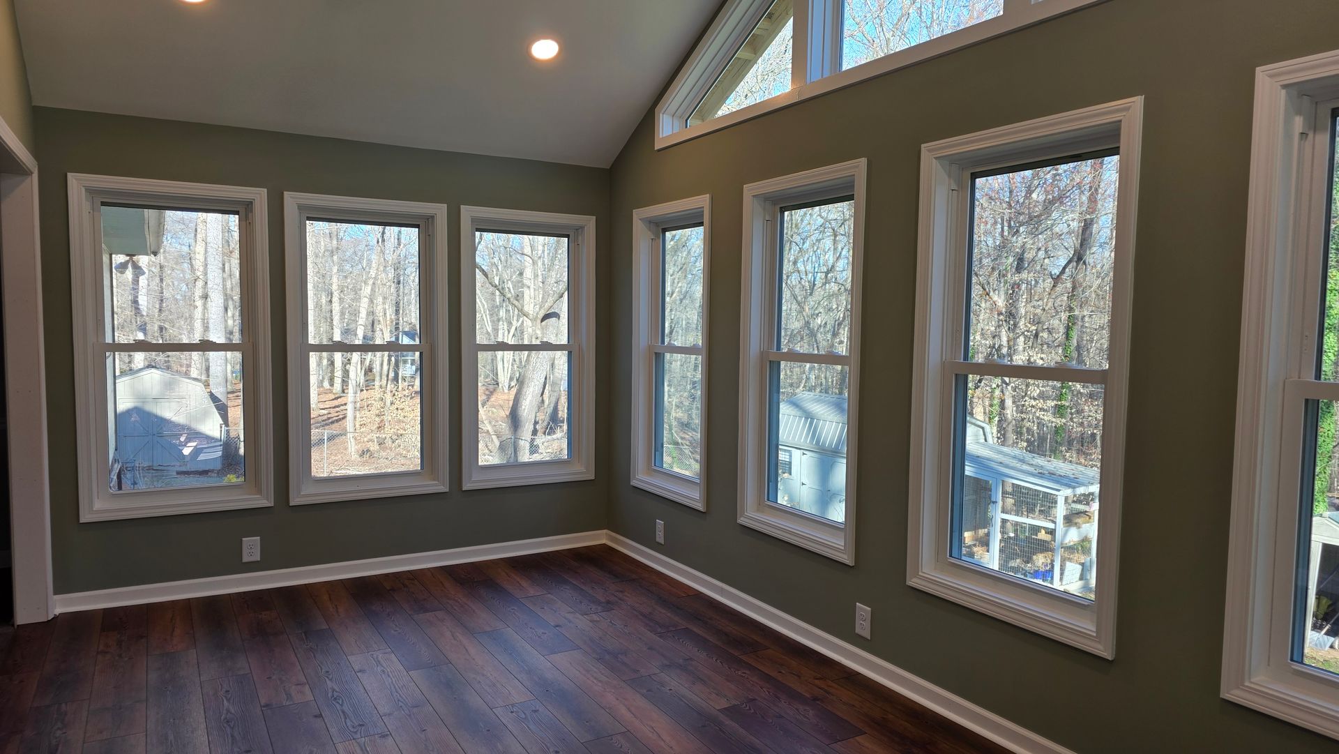 Sunroom interior with dark wood floors, olive green walls, and numerous windows offering a view of trees and a home exterior.