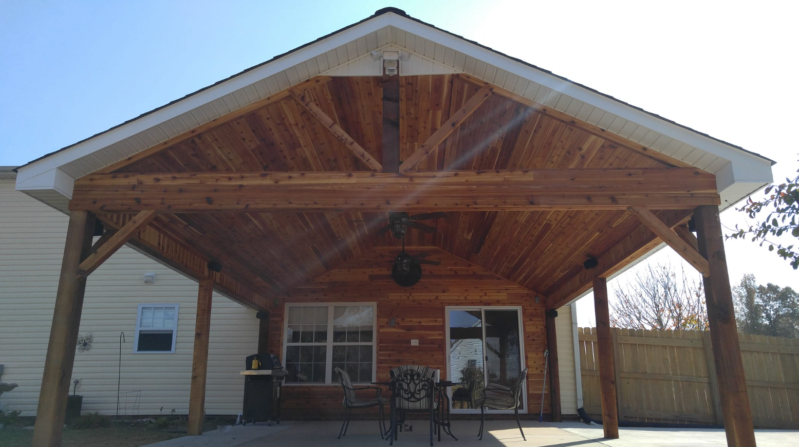 Wooden patio cover extending from a house, over a concrete patio with outdoor furniture. Sunny, clear day.