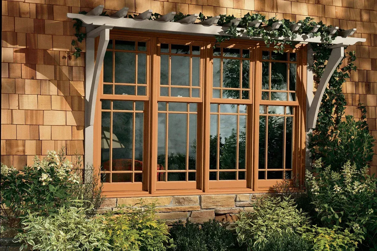 A wooden window with a trellis above, surrounded by greenery, set in a cedar shingled wall.