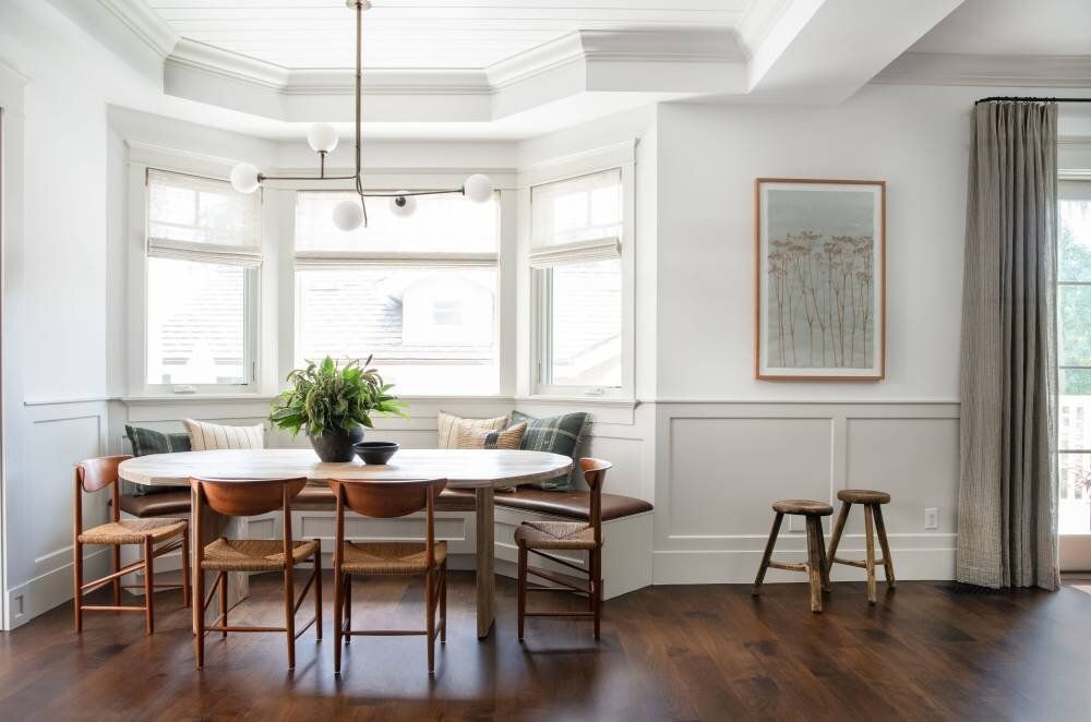 A bright dining room with a bay window and a table with woven chairs. A framed art piece hangs on the wall.