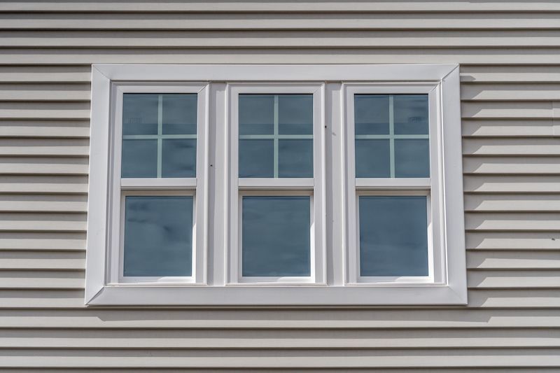 White-framed window with three panes on a light-colored, horizontally-sided wall. Each pane has a grid of white lines.